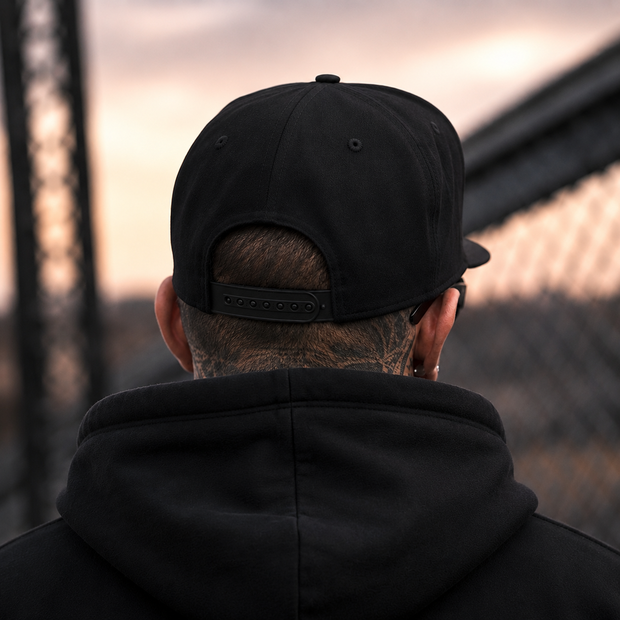 A man viewed from behind wearing a black snapback cap and a black hoodie, standing outdoors against a soft-focus urban background at sunset.