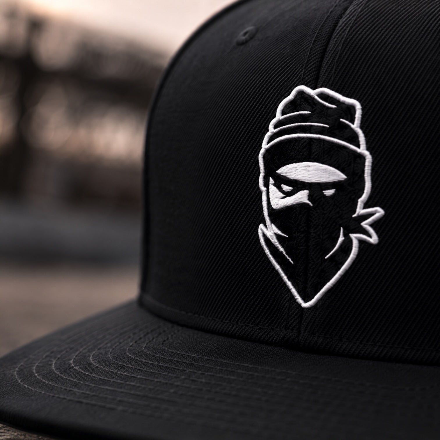 A close-up shot of a black snapback cap featuring a white embroidered logo of a ninja-like figure wearing a beanie and face mask, set against a blurred outdoor background.