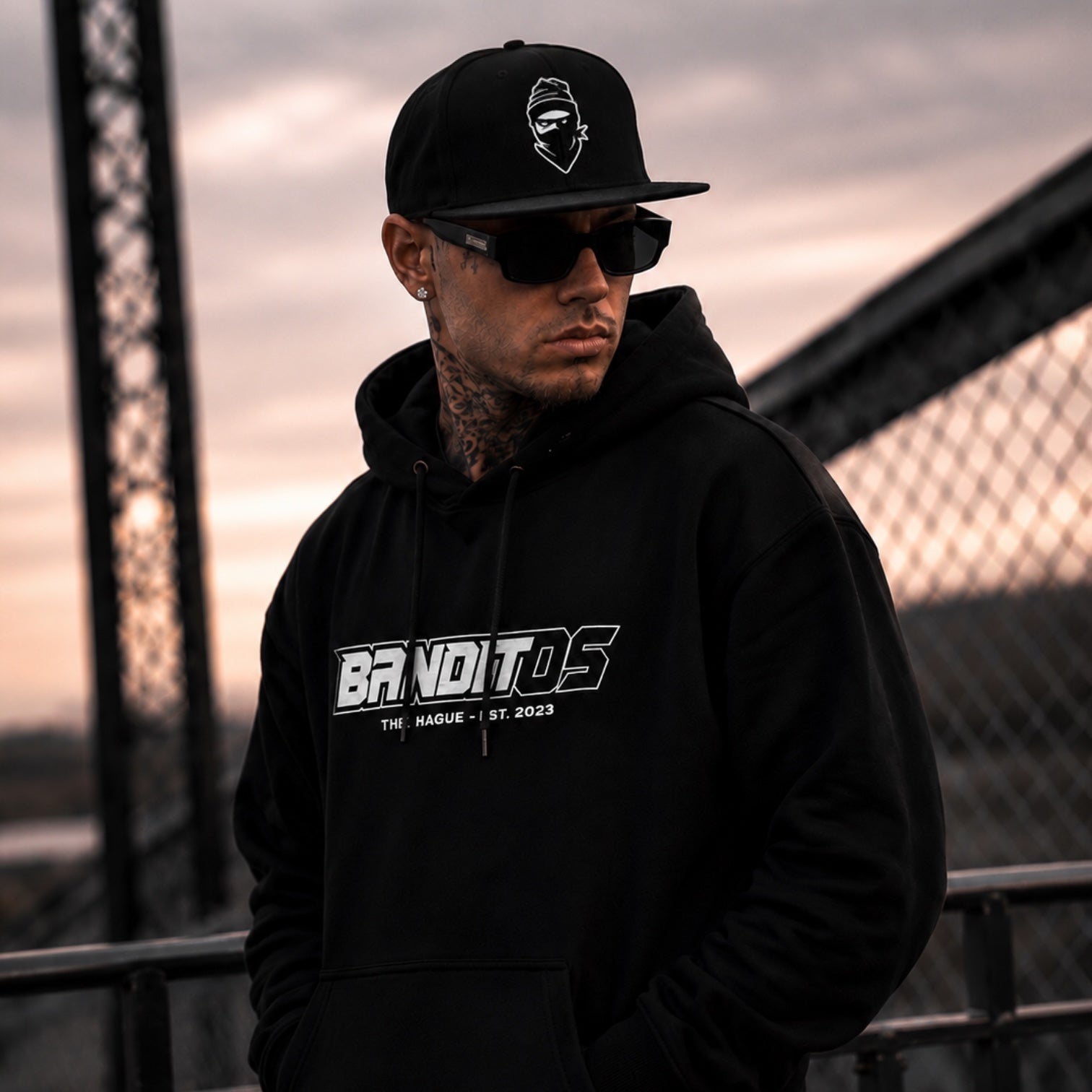 A man wearing a black hoodie and black cap stands outdoors in front of a bridge at sunset. The hoodie has a large white and gray 'BRANDITOS' graphic on the chest, and the cap features a small white logo.