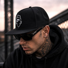 A man wearing a black snapback cap with a white graphic logo, black sunglasses, and a black hoodie, photographed outdoors against a blurred industrial background.