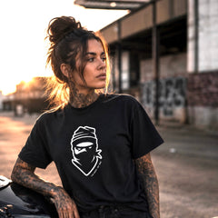 A woman with tattoos wearing a black graphic t-shirt with a white ninja face illustration, leaning against a car in an urban setting during sunset.