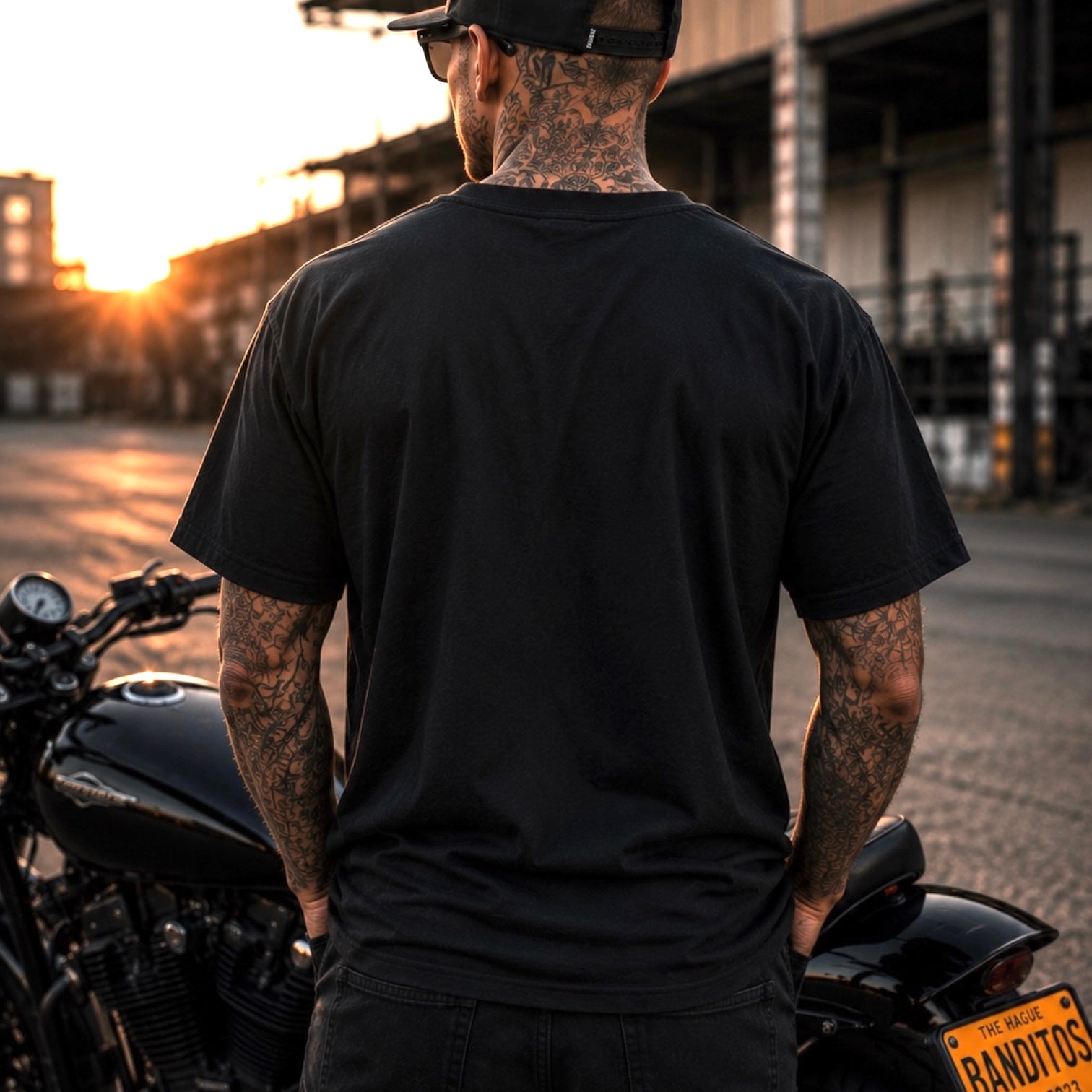 A man with tattoos on his neck and arms, wearing a black t-shirt and a black cap, standing in front of a black motorcycle at sunset in an industrial outdoor setting.