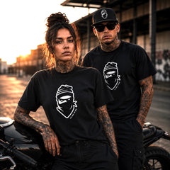 A man and a woman with visible tattoos wear matching black graphic T-shirts featuring a white ninja-style face logo, posed outdoors beside a motorcycle at sunset.