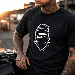 A man with tattooed arms wearing a black t-shirt with a white graphic of a masked figure, sitting on a motorcycle in an outdoor setting during sunset.