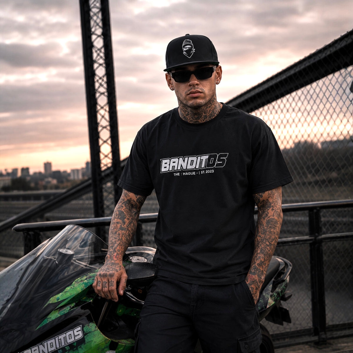 A man wearing a black BANDITOS t-shirt and black pants stands next to a green motorcycle on a bridge at sunset. The shirt features a prominent white and gray BANDITOS logo on the chest.