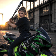 A woman in a black outfit sits on a black and neon green racing motorcycle, holding a black baseball bat. The setting is an industrial area with a warm sunset glow in the background.