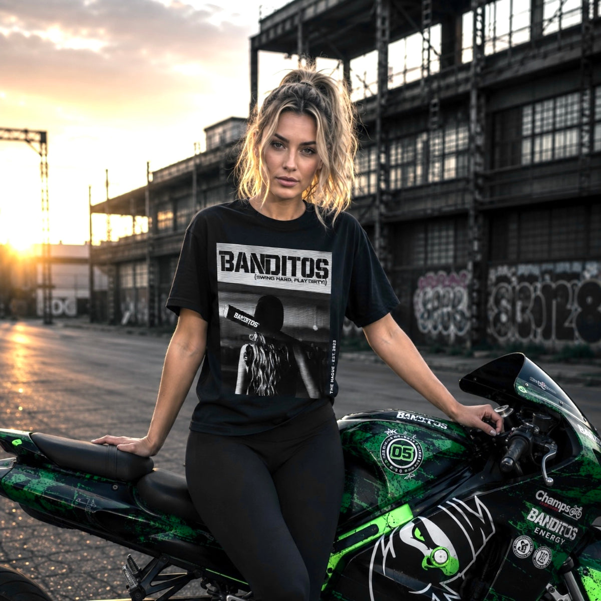 A woman posing on a green and black motorcycle in an industrial area at sunset, wearing a black graphic t-shirt with 'BANDITOS' and 'COSTUME PARTY' printed on the front.