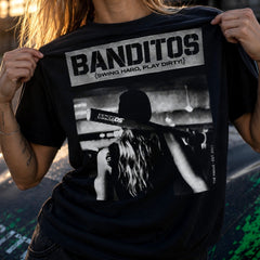 A person wearing a black graphic T-shirt with a large white and gray BANDITOS print on the front, holding the shirt open to show the design. The photo is taken outdoors with warm sunlight and a blurred street-like background.