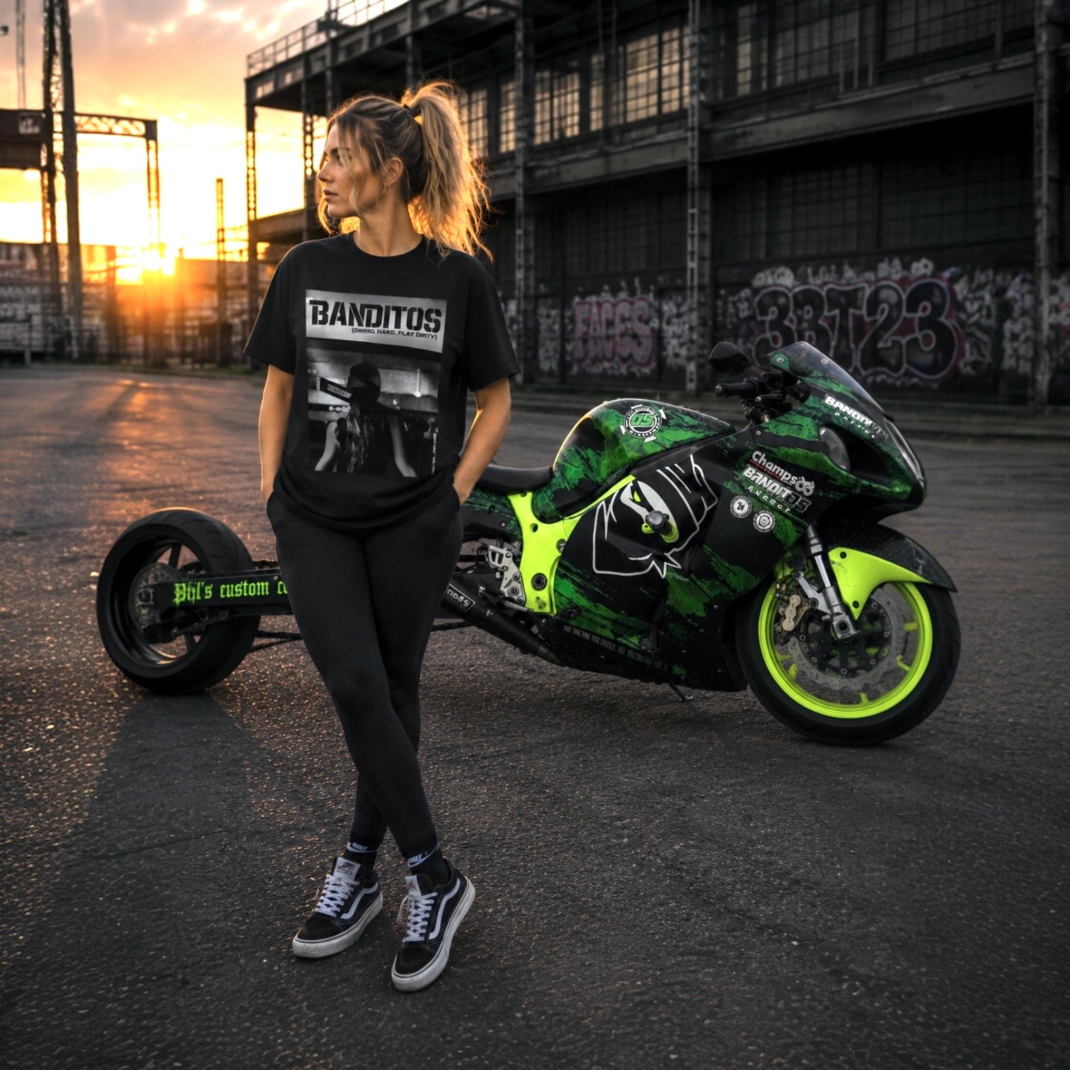 A woman stands outdoors in front of a green and black motorcycle, wearing a black graphic T-shirt with the word 'BANDITOS' and black leggings. The setting is a street at sunset with industrial buildings in the background.