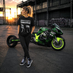 A woman stands outdoors in front of a green and black motorcycle, wearing a black graphic T-shirt with the word 'BANDITOS' and black leggings. The setting is a street at sunset with industrial buildings in the background.